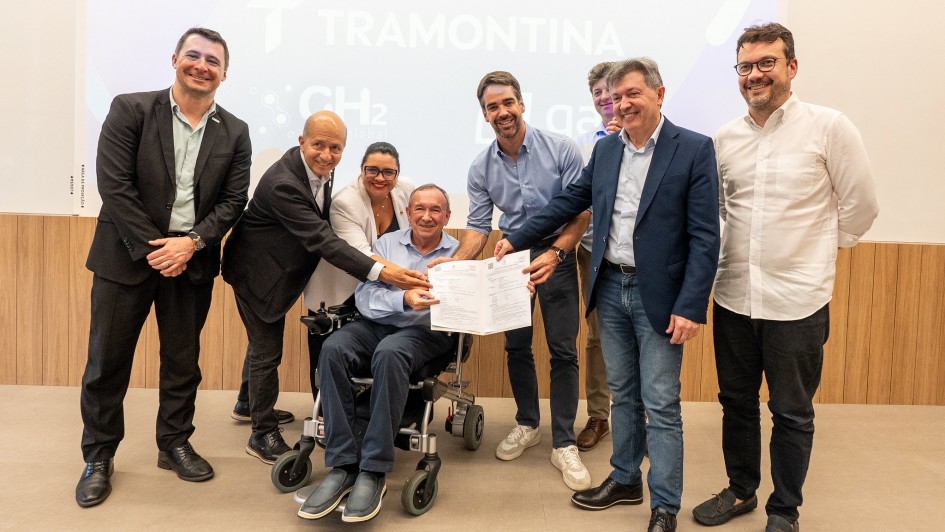 Foto posada de sete homens e uma mulher de corpo inteiro, exibindo um documento assinado e sorrindo para a câmera. Um dos homens estão numa cadeira de rodas, enquanto os outros estão de pé.