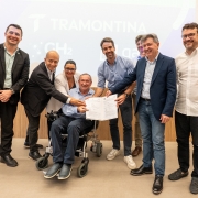 Foto posada de sete homens e uma mulher de corpo inteiro, exibindo um documento assinado e sorrindo para a câmera. Um dos homens estão numa cadeira de rodas, enquanto os outros estão de pé.