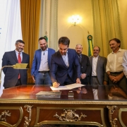 Foto de Leite assinando, de p&eacute;, um documento em uma mesa. Ao seu redor h&aacute; sete homens acompanhando a cena em ambiente formal.