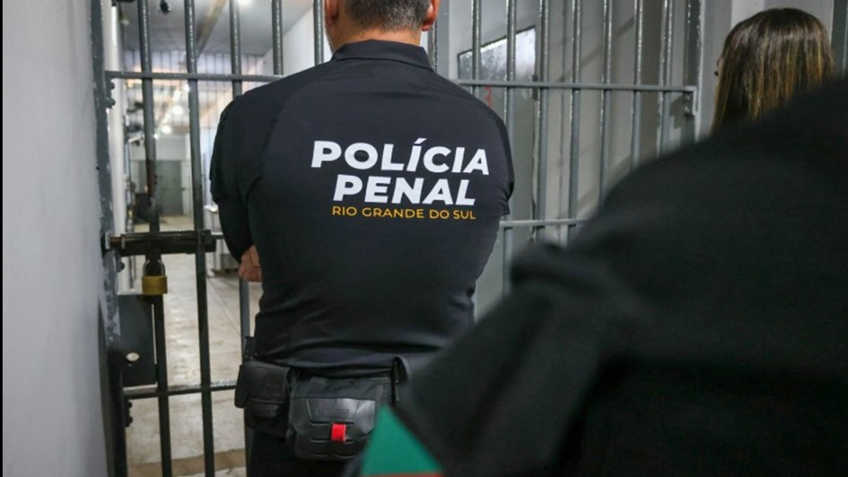 A imagem mostra um agente da Pol&iacute;cia Penal do Rio Grande do Sul, visto de costas. Ele est&aacute; diante de uma grade met&aacute;lica, t&iacute;pica de uma porta de cela, em um corredor interno de pres&iacute;dio. &Agrave; frente, h&aacute; outra pessoa parcialmente vis&iacute;vel, com um detalhe da bandeira do Rio Grande do Sul no bra&ccedil;o. O ambiente &eacute; fechado, com ilumina&ccedil;&atilde;o artificial e aspecto carcer&aacute;rio.