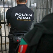 A imagem mostra um agente da Pol&iacute;cia Penal do Rio Grande do Sul, visto de costas. Ele est&aacute; diante de uma grade met&aacute;lica, t&iacute;pica de uma porta de cela, em um corredor interno de pres&iacute;dio. &Agrave; frente, h&aacute; outra pessoa parcialmente vis&iacute;vel, com um detalhe da bandeira do Rio Grande do Sul no bra&ccedil;o. O ambiente &eacute; fechado, com ilumina&ccedil;&atilde;o artificial e aspecto carcer&aacute;rio.