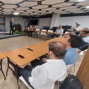 Foto mostra uma sala ampla, com um palestrante falando ao microfone ao fundo, e ao redor, sentados atr&aacute;s de mesas no formato U, est&atilde;o outros homens que acompanham a apresenta&ccedil;&atilde;o.