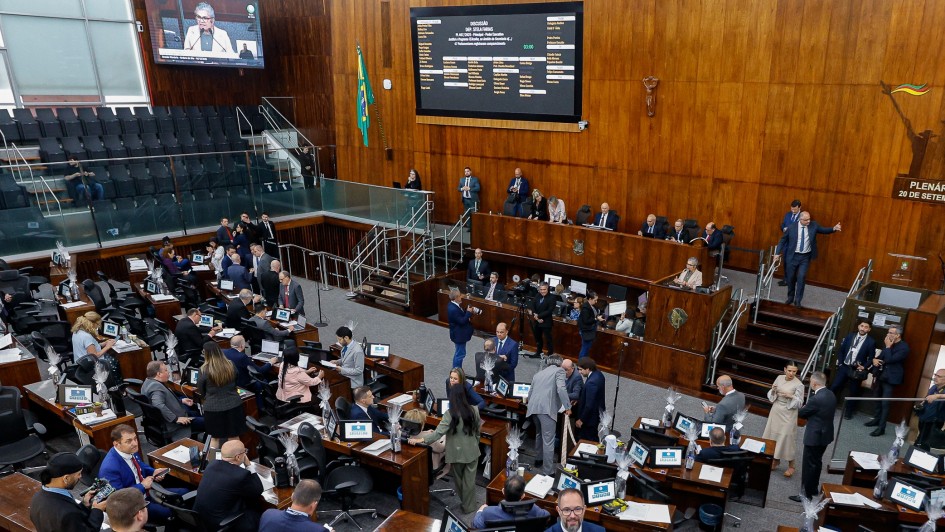 Uma fotografia panor&acirc;mica, tirada de um &acirc;ngulo superior e ligeiramente lateral, que mostra o interior de um Plen&aacute;rio da Assembleia Legislativa em sess&atilde;o.

O ambiente &eacute; amplo, com paredes e pain&eacute;is de madeira escura que revestem a maior parte das superf&iacute;cies verticais.

Centro e Base da Imagem: A &aacute;rea principal &eacute; ocupada pelas mesas e cadeiras (curules) dos deputados. H&aacute; uma grande movimenta&ccedil;&atilde;o, com muitos deputados e assessores em p&eacute; ou sentados, conversando e manuseando documentos ou laptops que est&atilde;o dispostos nas mesas de madeira escura.

Fundo da Imagem: Ao fundo, est&aacute; a Mesa Diretora, elevada em rela&ccedil;&atilde;o ao Plen&aacute;rio, com diversos membros sentados em cadeiras de encosto alto.

Telas: Duas telas grandes s&atilde;o vis&iacute;veis na parede de madeira. A tela &agrave; direita exibe uma lista de projetos ou vota&ccedil;&atilde;o, enquanto a tela &agrave; esquerda mostra a imagem de um orador.

Tribunas e Arquibancadas: &Agrave; esquerda, h&aacute; uma arquibancada com assentos pretos, onde algumas pessoas est&atilde;o sentadas e outras est&atilde;o em p&eacute;. Uma pessoa na parte superior segura uma faixa branca com letras vermelhas que diz "DESPES FICA!".

Decora&ccedil;&atilde;o: Na parede de madeira &agrave; direita, h&aacute; uma est&aacute;tua ou painel escultural de grandes dimens&otilde;es. A ilumina&ccedil;&atilde;o &eacute; forte, vinda de cima.

A cena sugere um momento de intensa atividade legislativa, provavelmente uma vota&ccedil;&atilde;o ou debate, dada a movimenta&ccedil;&atilde;o e a exibi&ccedil;&atilde;o de informa&ccedil;&otilde;es nas telas.