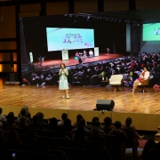 Secret&aacute;ria Raquel Teixeira est&aacute; num palco de piso de madeira polida. Ela fala em p&eacute; para uma plateia. Atr&aacute;s dela, um tel&atilde;o gigante reproduz a foto de outro evento, tamb&eacute;m em um audit&oacute;rio.
