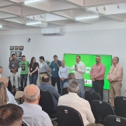 Foto mostra secret&aacute;rio falando em p&eacute; ao microfone, com prefeitos, lado a lado, enfileirados, acompanhando o discurso. Eles est&atilde;o diante de uma plateia sentada. Atr&aacute;s do palco h&aacute; um tel&atilde;o e bandeiras. 