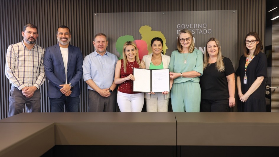 Foto mostra secret&aacute;ria e prefeito acompanhados de outras pessoas presentes no ato. A secret&aacute;ria e outras duas mulheres seguram um documento aberto.