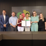 Foto mostra secret&aacute;ria e prefeito acompanhados de outras pessoas presentes no ato. A secret&aacute;ria e outras duas mulheres seguram um documento aberto.