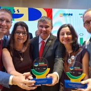 
Esta &eacute; uma fotografia em plano m&eacute;dio e close-up de cinco pessoas (tr&ecirc;s homens e duas mulheres) sorrindo e posando enquanto seguram dois trof&eacute;us em uma cerim&ocirc;nia de premia&ccedil;&atilde;o.

As Pessoas (da esquerda para a direita):

Homem 1: Veste terno azul, camisa branca e gravata bord&ocirc;. Ele usa &oacute;culos de grau e sorri abertamente.

Mulher 1: Tem cabelos castanhos longos, usa &oacute;culos de grau e um vestido texturizado em tom marrom escuro ou vinho. Ela usa um colar com pingente de cora&ccedil;&atilde;o dourado.

Homem 2 (ao centro): Um homem mais velho, vestindo terno preto, camisa branca e gravata vermelha. Ele est&aacute; posicionado no centro, com os bra&ccedil;os estendidos para segurar os dois trof&eacute;us junto com os colegas.

Mulher 2: Tem cabelos escuros e veste uma roupa escura com um detalhe bordado colorido (estilo &eacute;tnico ou tribal) no decote. Ela sorri diretamente para a c&acirc;mera.

Homem 3 (&agrave; direita): Veste uma camisa estampada azul com flores pequenas por baixo de um colete cinza. Ele tem barba curta, usa &oacute;culos e possui um pequeno brinco na orelha esquerda.

Os Trof&eacute;us: Eles seguram dois trof&eacute;us id&ecirc;nticos de design moderno.

A base &eacute; um ret&acirc;ngulo azul escuro com texto informativo em branco.

Sobre a base, h&aacute; um suporte em formato de semic&iacute;rculo (meia-lua) nas cores verde e amarelo.

A parte superior &eacute; um disco de acr&iacute;lico transparente onde se l&ecirc;: "PR&Ecirc;MIO BRASIL SEM FOME" em letras coloridas.

O trof&eacute;u da esquerda traz o texto na base: "BOM FUNCIONAMENTO DAS INST&Acirc;NCIAS DO SISAN".

O trof&eacute;u da direita traz: "REDU&Ccedil;&Atilde;O DA INSEGURAN&Ccedil;A ALIMENTAR E NUTRICIONAL NOS ESTADOS E NO DISTRITO FEDERAL".

O Fundo: Atr&aacute;s do grupo, h&aacute; uma grande tela de LED com gr&aacute;ficos vibrantes e abstratos em amarelo, vermelho, azul e verde. No canto superior direito da tela, &eacute; vis&iacute;vel o logotipo do SISAN e do Minist&eacute;rio do Desenvolvimento e Assist&ecirc;ncia Social, Fam&iacute;lia e Combate &agrave; Fome.

A cena &eacute; de celebra&ccedil;&atilde;o e vit&oacute;ria, com ilumina&ccedil;&atilde;o clara e foco centralizado no grupo e nos pr&ecirc;mios.