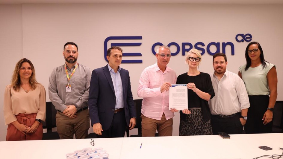 Esta &eacute; uma fotografia em plano m&eacute;dio de um grupo de sete pessoas (quatro homens e tr&ecirc;s mulheres) posando para uma foto em uma sala de reuni&otilde;es clara e moderna.

O Grupo (da esquerda para a direita):

Mulher 1: Possui cabelos castanhos longos, veste uma blusa bege de mangas compridas e cal&ccedil;as de cintura alta na cor terracota. Ela sorri suavemente com as m&atilde;os cruzadas &agrave; frente.

Homem 1: Tem barba curta e escura, veste uma camisa social cinza e usa um crach&aacute; pendurado em um cord&atilde;o colorido.

Homem 2: Veste um palet&oacute; azul-marinho sobre uma camisa social azul-claro, sem gravata.

Homem 3 (Centro): Veste uma camisa social rosa claro e cal&ccedil;as c&aacute;qui. Ele segura, junto com a mulher ao seu lado, uma folha de papel branca (um contrato ou conv&ecirc;nio).

Mulher 2 (Centro): Tem cabelos loiros curtos, usa &oacute;culos de arma&ccedil;&atilde;o preta, uma blusa preta e uma saia com estampa de leopardo (animal print). Ela segura a outra extremidade do documento.

Homem 4: Tem barba curta, veste uma camisa social branca e cal&ccedil;as pretas.

Mulher 3: Possui cabelos escuros longos, veste uma blusa verde-&aacute;gua clara sem mangas e cal&ccedil;as pretas.

O Fundo: Atr&aacute;s do grupo, h&aacute; uma parede branca e lisa. No centro da parede, acima das cabe&ccedil;as das pessoas, est&aacute; o logotipo da CORSAN (Companhia Riograndense de Saneamento) em azul escuro, composto por tr&ecirc;s barras horizontais que formam um "C" estilizado, seguido pelo nome "corsan" e um pequeno s&iacute;mbolo "ae" no canto superior direito.

O Primeiro Plano: Na parte inferior da imagem, v&ecirc;-se parte de uma grande mesa de reuni&otilde;es branca. Sobre ela, h&aacute; uma bandeja com diversos copos pl&aacute;sticos lacrados contendo &aacute;gua, al&eacute;m de alguns cabos e um pequeno dispositivo eletr&ocirc;nico preto no canto direito.

A ilumina&ccedil;&atilde;o &eacute; uniforme e clara, vindo de pain&eacute;is de LED no teto, criando um ambiente corporativo formal.