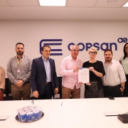 Esta &eacute; uma fotografia em plano m&eacute;dio de um grupo de sete pessoas (quatro homens e tr&ecirc;s mulheres) posando para uma foto em uma sala de reuni&otilde;es clara e moderna.

O Grupo (da esquerda para a direita):

Mulher 1: Possui cabelos castanhos longos, veste uma blusa bege de mangas compridas e cal&ccedil;as de cintura alta na cor terracota. Ela sorri suavemente com as m&atilde;os cruzadas &agrave; frente.

Homem 1: Tem barba curta e escura, veste uma camisa social cinza e usa um crach&aacute; pendurado em um cord&atilde;o colorido.

Homem 2: Veste um palet&oacute; azul-marinho sobre uma camisa social azul-claro, sem gravata.

Homem 3 (Centro): Veste uma camisa social rosa claro e cal&ccedil;as c&aacute;qui. Ele segura, junto com a mulher ao seu lado, uma folha de papel branca (um contrato ou conv&ecirc;nio).

Mulher 2 (Centro): Tem cabelos loiros curtos, usa &oacute;culos de arma&ccedil;&atilde;o preta, uma blusa preta e uma saia com estampa de leopardo (animal print). Ela segura a outra extremidade do documento.

Homem 4: Tem barba curta, veste uma camisa social branca e cal&ccedil;as pretas.

Mulher 3: Possui cabelos escuros longos, veste uma blusa verde-&aacute;gua clara sem mangas e cal&ccedil;as pretas.

O Fundo: Atr&aacute;s do grupo, h&aacute; uma parede branca e lisa. No centro da parede, acima das cabe&ccedil;as das pessoas, est&aacute; o logotipo da CORSAN (Companhia Riograndense de Saneamento) em azul escuro, composto por tr&ecirc;s barras horizontais que formam um "C" estilizado, seguido pelo nome "corsan" e um pequeno s&iacute;mbolo "ae" no canto superior direito.

O Primeiro Plano: Na parte inferior da imagem, v&ecirc;-se parte de uma grande mesa de reuni&otilde;es branca. Sobre ela, h&aacute; uma bandeja com diversos copos pl&aacute;sticos lacrados contendo &aacute;gua, al&eacute;m de alguns cabos e um pequeno dispositivo eletr&ocirc;nico preto no canto direito.

A ilumina&ccedil;&atilde;o &eacute; uniforme e clara, vindo de pain&eacute;is de LED no teto, criando um ambiente corporativo formal.