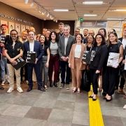
Esta &eacute; uma fotografia em plano geral de um grupo numeroso de pessoas (cerca de 25 a 30 indiv&iacute;duos) posando para uma foto oficial em um ambiente interno institucional.

O Grupo:

Composto por homens e mulheres de diversas idades, vestidos predominantemente com trajes casuais e profissionais (blazers, camisas, blusas e cal&ccedil;as sociais).

Est&atilde;o distribu&iacute;dos de p&eacute;, ocupando o centro e as laterais do quadro.

Pelo menos cinco pessoas, distribu&iacute;das pelo grupo, seguram &agrave; frente do corpo uma pasta ou livreto preto com a palavra "CULTURA" escrita verticalmente em letras brancas grandes e em negrito.

A maioria das pessoas est&aacute; sorrindo, voltada para a c&acirc;mera.

O Ambiente:

&Agrave; esquerda: H&aacute; uma parede marrom escura com a inscri&ccedil;&atilde;o em letras met&aacute;licas: "GALERIA DOS EX-SECRET&Aacute;RIOS DA CULTURA". Abaixo do texto, h&aacute; v&aacute;rias molduras com retratos fotogr&aacute;ficos de antigos gestores alinhados em duas fileiras.

Ao fundo: No centro, acima de um corredor, h&aacute; uma placa de sinaliza&ccedil;&atilde;o cinza que indica: "SECRETARIA DA CULTURA SEDAC". A ilumina&ccedil;&atilde;o do teto &eacute; feita por trilhos com spots direcionais brancos.

&Agrave; direita: Parte de uma parede exibe um cartaz ou obra de arte emoldurada com elementos gr&aacute;ficos coloridos.

O ch&atilde;o: Revestido com grandes lajetas de cer&acirc;mica cinza. No lado direito, destaca-se uma faixa de piso t&aacute;til de alerta na cor amarela, que atravessa verticalmente parte do cen&aacute;rio.

A imagem registra um encontro ou evento oficial na sede da Secretaria de Estado da Cultura (Sedac) do Rio Grande do Sul.