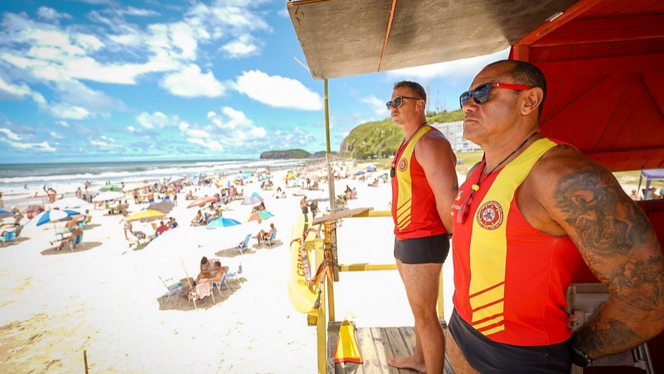 Foto de lado de dois guarda-vidas de p&eacute; na guarita, olhando para o mar, que aparece &agrave; esquerda da foto. O dia est&aacute; ensolarado e com v&aacute;rias barracas e pessoas na praia.