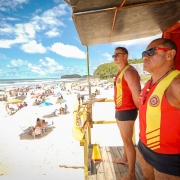 Foto de lado de dois guarda-vidas de p&eacute; na guarita, olhando para o mar, que aparece &agrave; esquerda da foto. O dia est&aacute; ensolarado e com v&aacute;rias barracas e pessoas na praia.
