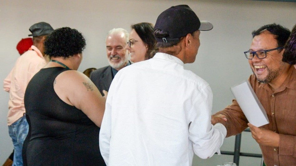 Foto mostra alunos do curso recebendo os certificados. Eles est&atilde;o de costas para a foto e de frente para os respons&aacute;veis pelo curso que entregam os certificados. Em destaque, um deles, de bon&eacute; preto e camisa branca, &eacute; cumprimentado por um homem de &oacute;culos que o sa&uacute;da com um aperto de m&atilde;o e est&aacute; muito sorridente.