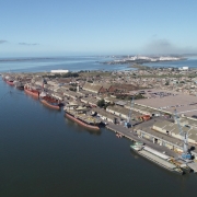 Imagem a&eacute;rea de um porto p&uacute;blico &agrave;s margens de um rio largo e calmo. V&aacute;rios navios de carga de grande porte est&atilde;o atracados em sequ&ecirc;ncia ao longo dos cais. &Agrave; direita, h&aacute; extensos armaz&eacute;ns portu&aacute;rios, guindastes e &aacute;reas pavimentadas para opera&ccedil;&atilde;o log&iacute;stica. &Agrave; esquerda do rio, aparecem &aacute;reas de vegeta&ccedil;&atilde;o baixa e ilhas fluviais. Ao fundo, observa-se a foz e a continuidade do curso d&rsquo;&aacute;gua em dire&ccedil;&atilde;o ao horizonte, sob c&eacute;u azul claro.