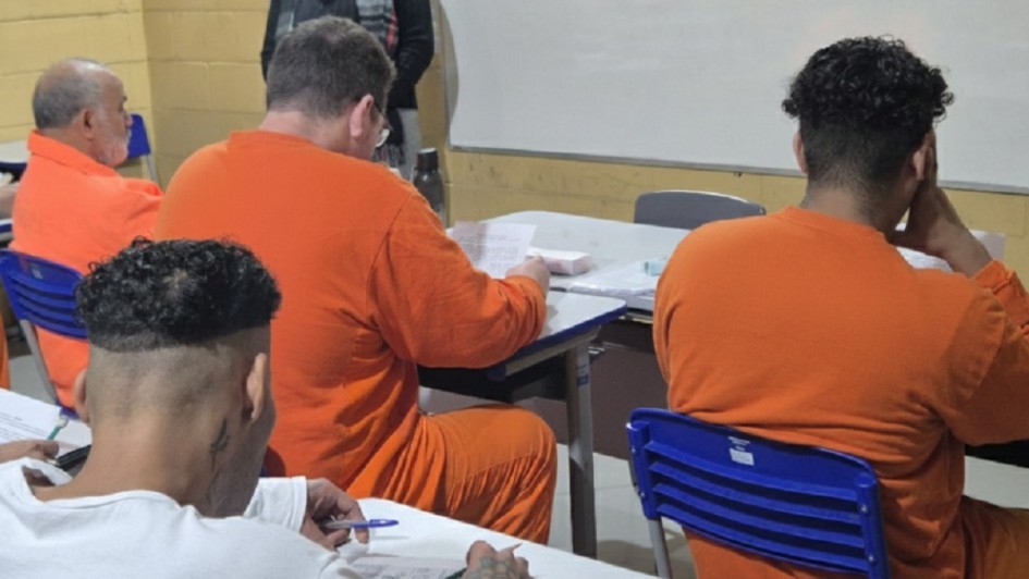 Foto mostra quatro apenados sentados em carteiras de escola. Eles est&atilde;o de costas para a foto. Tr&ecirc;s deles vestem uniformes laranjas.