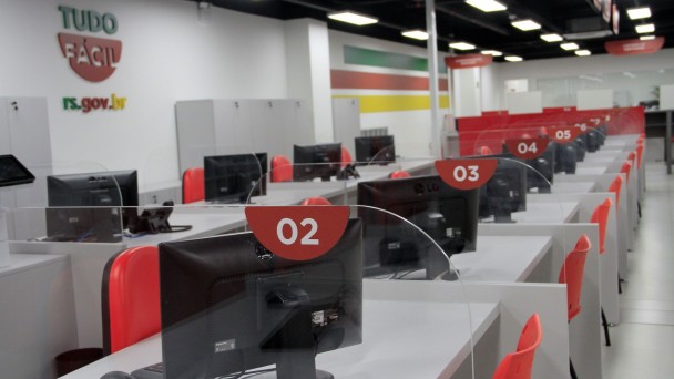 Imagem mostra uma fileira de guich&ecirc;s do Tudo F&aacute;cil de Passo Fundo, com computadores sobre as mesas cinzas, cadeiras laranjas e identifica&ccedil;&atilde;o com os n&uacute;meros dos guich&ecirc;s em placas vermelhas com n&uacute;meros brancos. Ao fundo parede branca com s&iacute;mbolo do Tudo F&aacute;cil.