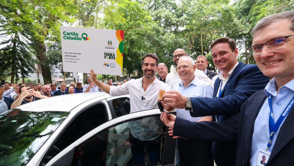 Foto ao ar livre posada, de Leite segurando uma grande r&eacute;plica de um cart&atilde;o cidad&atilde;o nas m&atilde;os, enquanto sorri. Ele est&aacute; ao lado de um dos ve&iacute;culos entregues. H&aacute; quatro homens junto a ele no primeiro plano, e v&aacute;rias ao fundo, num ambiente arborizado.