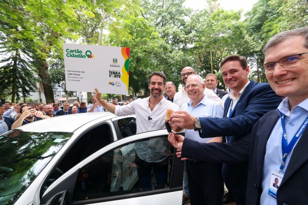 Foto ao ar livre posada, de Leite segurando uma grande réplica de um cartão cidadão nas mãos, enquanto sorri. Ele está ao lado de um dos veículos entregues. Há quatro homens junto a ele no primeiro plano, e várias ao fundo, num ambiente arborizado.