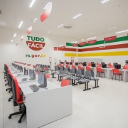 Foto mostra imagem da sala da ag&ecirc;ncia, com muitos guich&ecirc;s na cor cinza claro e cadeiras laranjas e sobre eles computadores. Ao fundo, a logomarca do Tudo F&aacute;cil e listas amarelas vermelhas e verdes decorando as paredes. 