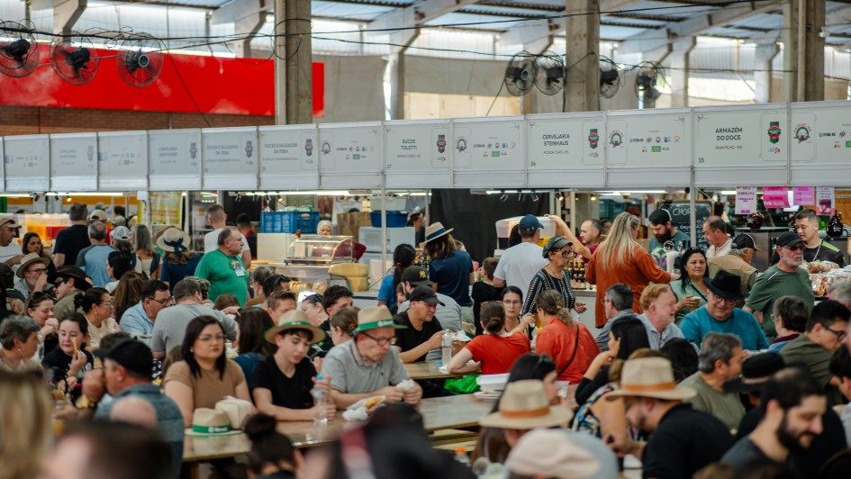 A imagem mostra o Pavilh&atilde;o da Agricultura Familiar na Expointer 2025. O local est&aacute; cheio de pessoas e h&aacute; diversas bancas de produtos variados. 