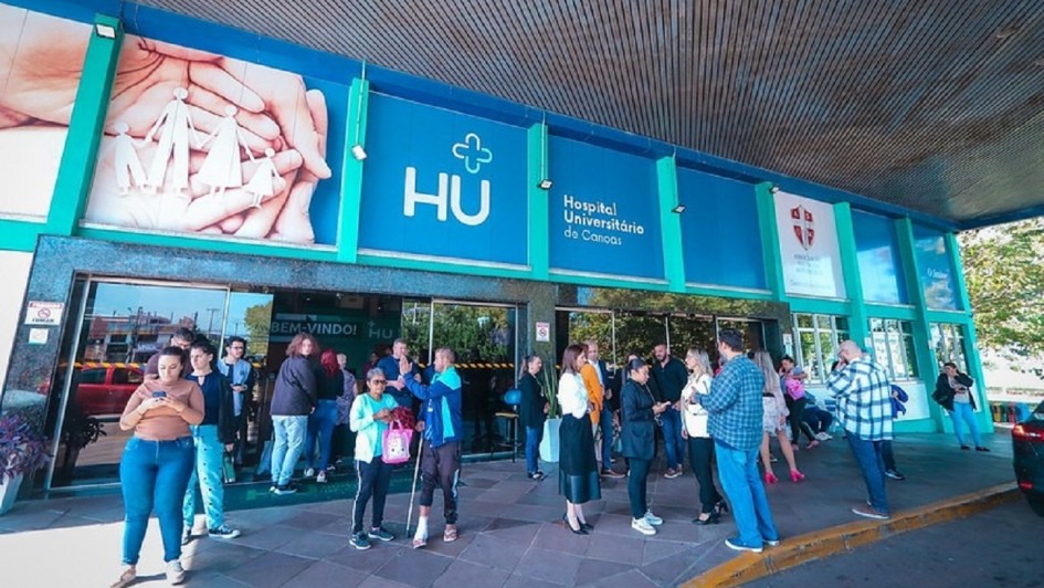 Esta &eacute; uma descri&ccedil;&atilde;o da imagem para fins de acessibilidade:

A fotografia mostra a fachada e a entrada principal do Hospital Universit&aacute;rio de Canoas (HU) em um dia claro e ensolarado.

Detalhes da Fachada
O pr&eacute;dio apresenta uma estrutura moderna com pain&eacute;is em tons de azul vibrante e verde.

&Agrave; esquerda, h&aacute; um grande painel art&iacute;stico mostrando m&atilde;os que protegem a silhueta de uma fam&iacute;lia (pai, m&atilde;e e duas crian&ccedil;as).

Ao centro, destaca-se o logotipo do hospital: uma cruz estilizada acima das letras grandes "HU", seguidas pelo texto "Hospital Universit&aacute;rio de Canoas".

&Agrave; direita, h&aacute; outros logotipos menores, incluindo o da associa&ccedil;&atilde;o que faz a gest&atilde;o do local.

Elementos da Cena
Pessoas: Cerca de vinte pessoas est&atilde;o distribu&iacute;das em frente &agrave; entrada. Algumas conversam em grupos, outras est&atilde;o sozinhas olhando o celular ou caminhando.

Destaque: Pr&oacute;ximo ao centro da imagem, um homem utiliza uma muleta e est&aacute; com o p&eacute; esquerdo enfaixado, conversando com uma mulher de blusa verde.

Ambiente: A cal&ccedil;ada &eacute; feita de pedras irregulares em tons de cinza e marrom. A entrada principal possui portas de vidro escuro que refletem o movimento externo.