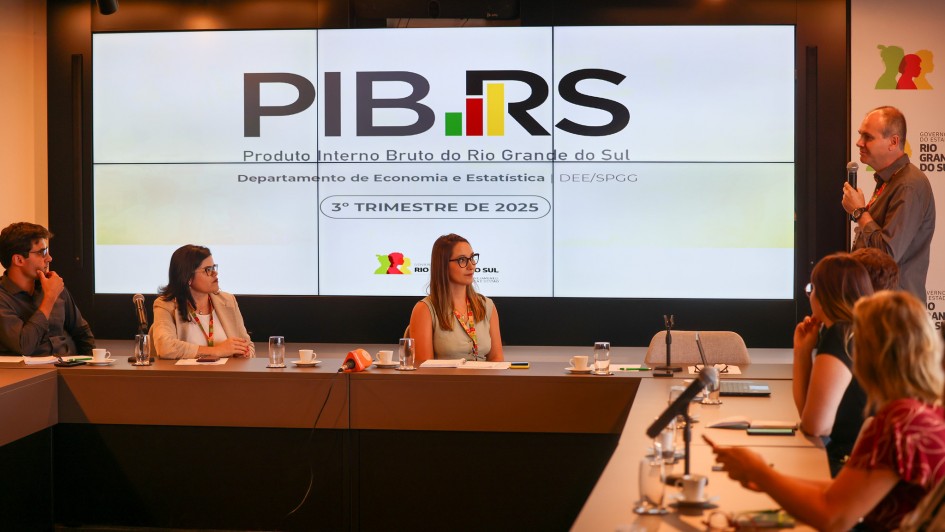 A imagem mostra uma mesa retangular com diversas pessoas sentadas ao redor. No canto direito, h&aacute; um homem em p&eacute; falando ao microfone. No painel ao fundo, l&ecirc;-se "PIB RS" em destaque. 