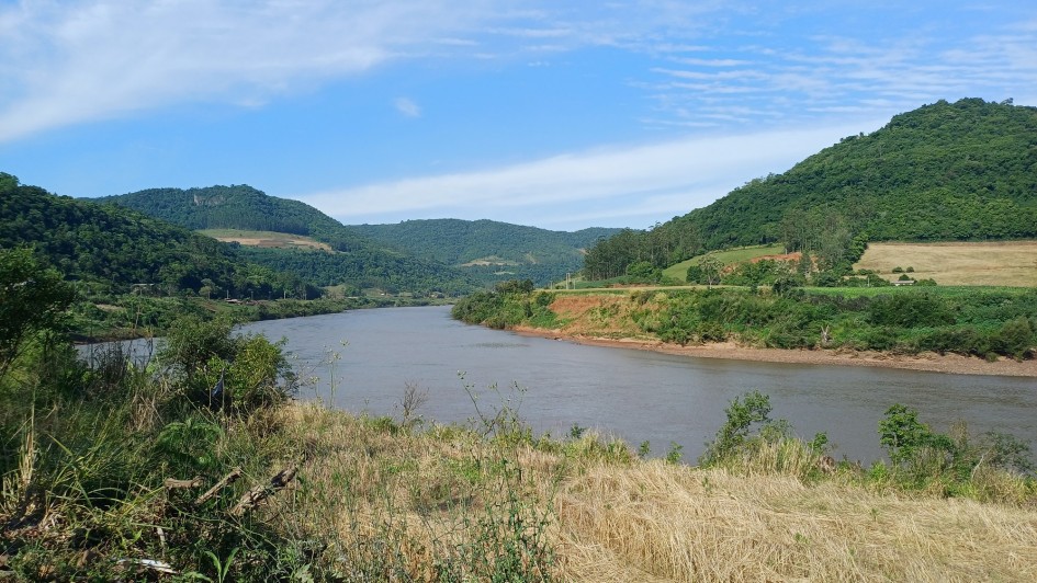 A fotografia apresenta uma vista panor&acirc;mica de um rio largo que serpenteia por um vale entre colinas cobertas de vegeta&ccedil;&atilde;o, sob um c&eacute;u azul claro.

Detalhes da Paisagem
O Rio: O curso d'&aacute;gua ocupa a parte central da imagem, apresentando &aacute;guas de tom acastanhado e uma superf&iacute;cie relativamente calma que reflete a claridade do dia.

O Relevo: Ao fundo e nas laterais, elevam-se morros e colinas cobertos por uma mata verde e densa. Algumas &aacute;reas nas encostas mostram clar&otilde;es de terra e campos cultivados, evidenciando o uso agr&iacute;cola da terra na regi&atilde;o.

Primeiro Plano: Na parte inferior da foto, v&ecirc;-se a vegeta&ccedil;&atilde;o da margem, composta por arbustos verdes, gram&iacute;neas secas de cor amarelada e um solo pedregoso no canto esquerdo.

O C&eacute;u: O c&eacute;u est&aacute; bem aberto e azul, com nuvens brancas finas e alongadas que se espalham de forma suave.

Contexto
De acordo com o t&iacute;tulo do arquivo, a imagem ilustra a implementa&ccedil;&atilde;o de um novo servi&ccedil;o de monitoramento pelo Governo do Estado do Rio Grande do Sul, projetado para prever o n&iacute;vel dos rios e auxiliar na preven&ccedil;&atilde;o de enchentes.