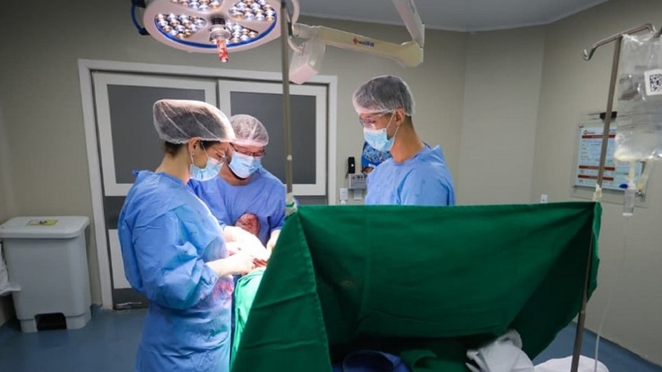 Tr&ecirc;s profissionais de sa&uacute;de realizam cirurgia em sala de opera&ccedil;&atilde;o, com equipamentos m&eacute;dicos e ilumina&ccedil;&atilde;o intensa.