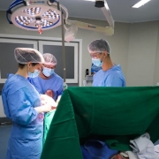 Tr&ecirc;s profissionais de sa&uacute;de realizam cirurgia em sala de opera&ccedil;&atilde;o, com equipamentos m&eacute;dicos e ilumina&ccedil;&atilde;o intensa.