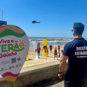 A imagem mostra um homem de uniforme da Receita Estadual de costas. Ao fundo, est&aacute; o mar e banhistas. H&aacute; uma placa no lado esquerdo onde se l&ecirc; "Viva o ver&atilde;o ga&uacute;cho". 