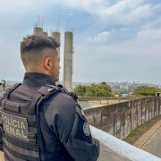 Esta &eacute; uma descri&ccedil;&atilde;o detalhada da imagem para fins de acessibilidade:

A imagem apresenta um policial penal visto de costas e de perfil, em um plano m&eacute;dio. Ele observa o horizonte a partir de um ponto elevado, possivelmente uma guarita ou o topo de um muro de uma unidade prisional.

Detalhes do Policial:
Vestimenta: Ele utiliza um uniforme operacional preto e um colete bal&iacute;stico da mesma cor. Nas costas do colete, h&aacute; um adesivo retangular cinza com a inscri&ccedil;&atilde;o "POL&Iacute;CIA PENAL" em letras mai&uacute;sculas pretas.

Identifica&ccedil;&atilde;o: No bra&ccedil;o direito, h&aacute; um patch (emblema) circular preto com detalhes em branco, onde se l&ecirc; "PEPOA" (provavelmente referente a um grupamento espec&iacute;fico).

Apar&ecirc;ncia: O homem tem pele clara, cabelo castanho escuro com corte curto nas laterais (estilo undercut) e barba aparada.

Cen&aacute;rio e Composi&ccedil;&atilde;o:
Primeiro Plano: O policial est&aacute; posicionado &agrave; esquerda, apoiado ou pr&oacute;ximo a um parapeito de concreto claro. &Agrave; sua frente, v&ecirc;-se um muro alto com grafites coloridos e desgastados.

Segundo Plano: H&aacute; grandes estruturas cil&iacute;ndricas cinzas que se assemelham a caixas d'&aacute;gua industriais ou silos. &Agrave; direita, uma rua asfaltada com um carro branco estacionado.

Fundo: Ao longe, &eacute; poss&iacute;vel ver a silhueta de uma &aacute;rea urbana densamente povoada, com diversos pr&eacute;dios sob um c&eacute;u claro com nuvens esparsas.

A ilumina&ccedil;&atilde;o &eacute; de luz do dia natural, sugerindo um clima aberto e ensolarado. A composi&ccedil;&atilde;o transmite uma sensa&ccedil;&atilde;o de vigil&acirc;ncia e monitoramento.