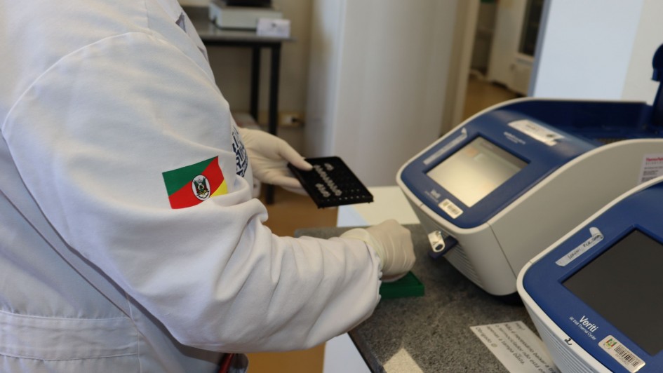 A imagem mostra uma pessoa de jaleco branco e luvas trabalhando em uma bancada, em um ambiente laboratorial. 