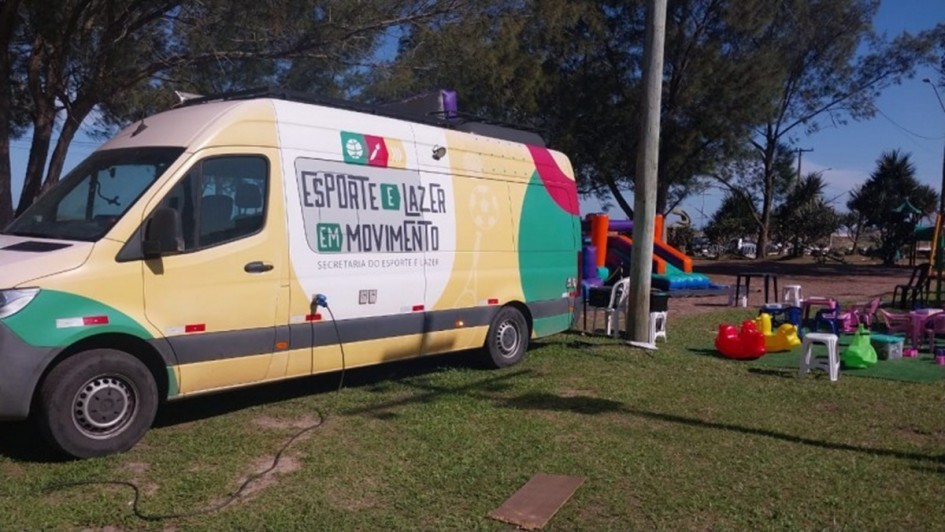 Foto mostra van do programa com adesivagem colorida e alguns brinquedos infantis pr&oacute;ximos. O ve&iacute;culo est&aacute; estacionado em um gramado, com uma &aacute;rvore grande atr&aacute;s.