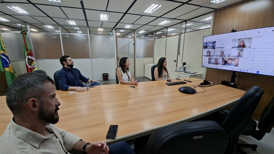 A imagem mostra uma mesa retangular com pessoas reunidas em um ambiente de trabalho. No canto direito, h&aacute; uma tela com pessoas participando de forma remota da reuni&atilde;o. 