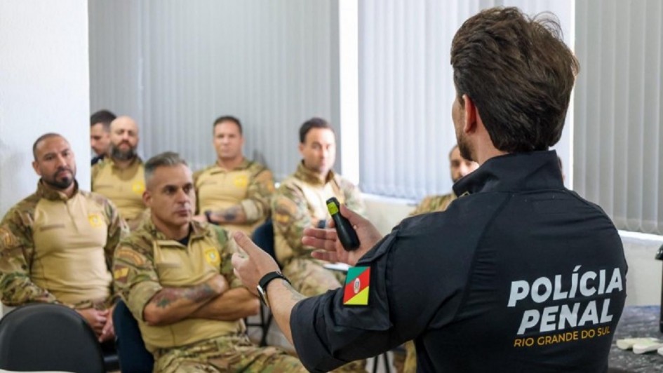 Foto mostra instrutor da Pol&iacute;cia Penal, de camisa preta com o nome da institui&ccedil;&atilde;o, falando para outros colegas fardados, de farda bege clara. 