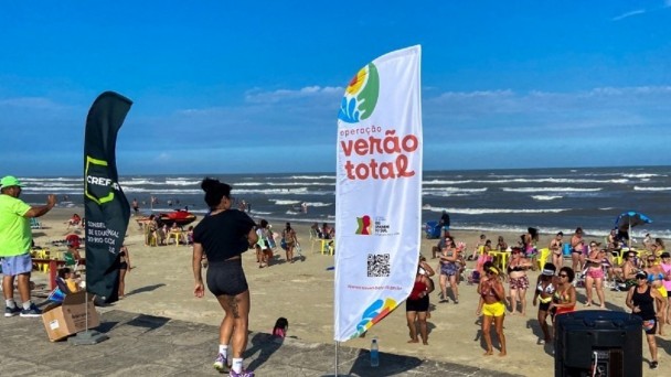 Foto mostra instrutora dando aula de dança na beira da praia. O mar está ao fundo. As aulas estão na areia. Há um banner da Operação Verão Total.
