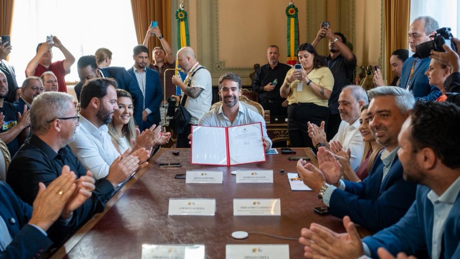 Governador Eduardo Leite segura documento. Ele est&aacute; sentado na ponta de uma mesa onde h&aacute; outras autoridades que batem palmas. Ao fundo, v&aacute;rios assessores est&atilde;o tirando foto do momento.