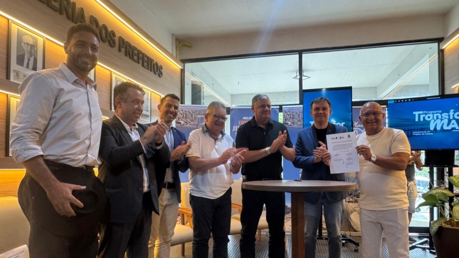 Uma fotografia colorida em plano m&eacute;dio mostra um grupo de sete homens em p&eacute;, comemorando a assinatura de um documento em um ambiente interno.

A&ccedil;&atilde;o Principal: O grupo est&aacute; posicionado em semic&iacute;rculo atr&aacute;s de uma pequena mesa redonda de madeira. O homem na extremidade direita, vestindo camiseta branca e cal&ccedil;a clara, segura um documento assinado com as duas m&atilde;os e sorri para a c&acirc;mera. Os outros seis homens ao seu lado est&atilde;o aplaudindo.

Os Participantes:

&Agrave; esquerda, o primeiro homem veste camisa branca e cal&ccedil;a escura, segurando um chap&eacute;u preto na m&atilde;o.

Os demais vestem roupas sociais ou casuais elegantes, incluindo palet&oacute;s, camisas polo e camisas sociais em tons de azul, branco e preto.

O Ambiente:

Ao fundo, na parede esquerda, h&aacute; uma placa dourada com os dizeres "GALERIA DOS PREFEITOS", acima de v&aacute;rios quadros com fotos em preto e branco.

No centro e &agrave; direita, h&aacute; telas de TV exibindo a identidade visual do "Programa Transforma Tramanda&iacute;".

O ch&atilde;o possui um tapete de fios longos e claros sob a mesa, e o restante do piso &eacute; escuro e texturizado.

Contexto: A cena sugere um ato oficial de assinatura de conv&ecirc;nio ou financiamento entre o governo e o munic&iacute;pio.