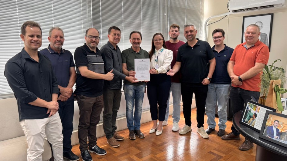Uma fotografia colorida em plano m&eacute;dio-longo mostra um grupo de 11 pessoas (dez homens e uma mulher) posando para uma foto oficial em um gabinete ou escrit&oacute;rio.

A&ccedil;&atilde;o Central: No centro do grupo, uma mulher e um homem seguram juntos um documento oficial em papel branco, exibindo-o para a c&acirc;mera como o registro de uma assinatura ou acordo.

Os Participantes: As pessoas est&atilde;o alinhadas lado a lado, formando uma fila levemente curvada. Elas vestem roupas de estilo casual e casual-fino, como camisas polo, camisas sociais de mangas curtas e compridas, camisetas e cal&ccedil;as jeans ou de sarja. A maioria sorri de forma amig&aacute;vel.

O Ambiente:

O ch&atilde;o &eacute; revestido com um piso de madeira (parquet) em tons de mel.

Ao fundo, h&aacute; uma grande janela coberta por persianas horizontais brancas fechadas.

No canto superior direito, v&ecirc;-se um aparelho de ar-condicionado do tipo split.

No canto inferior direito, h&aacute; uma mesa de apoio com dois porta-retratos (um deles com a foto do governador Eduardo Leite), uma planta ornamental e uma sacola de presente parda com la&ccedil;o verde.

No canto inferior esquerdo, aparece uma parte de um sof&aacute; ou poltrona de tecido claro.