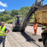 A imagem mostra obras em uma rodovia, com trabalhadores e máquinas na pista. O dia está ensolarado.