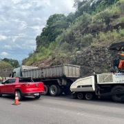A imagem mostra uma rodovia em obras, com máquinas, carros e trabalhadores na pista.