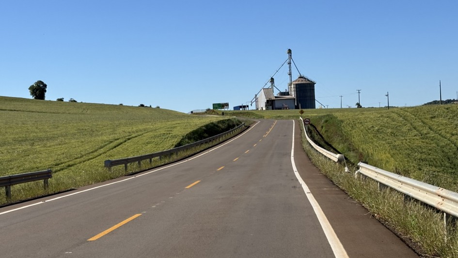 Foto mostra uma estrada ao centro, com campos verdes nas laterais e um silo de gr&atilde;os ao fim de estrada, em menor perspectiva. O c&eacute;u est&aacute; azul, sem nuvens.