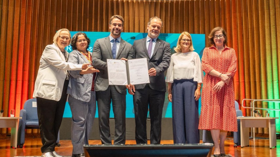 Seis pessoas est&atilde;o em p&eacute; em um palco, posando para a foto e segurando um documento assinado. 