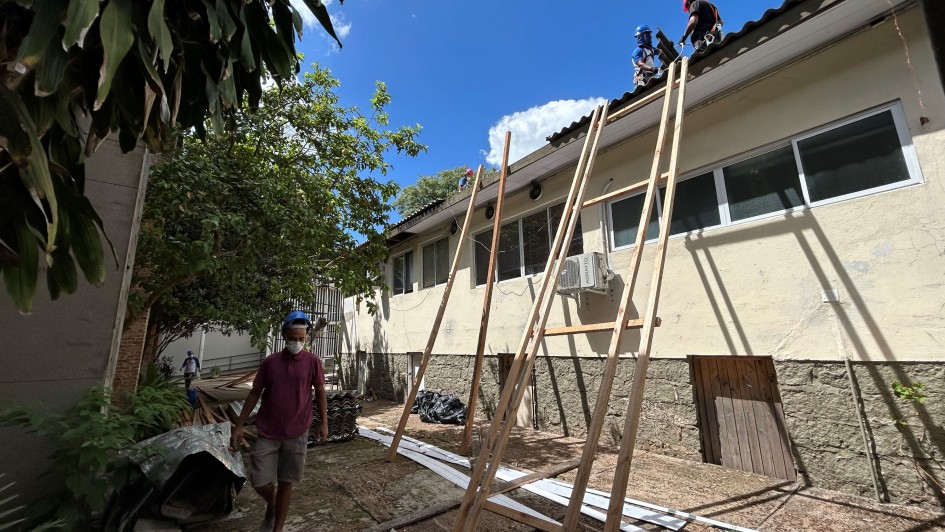 Uma fotografia colorida em plano aberto captura o trabalho de reforma no telhado de um pr&eacute;dio de dois andares, identificado como o setor de Tisiologia do Sanat&oacute;rio Partenon.

A&ccedil;&atilde;o Principal: No topo do pr&eacute;dio, &agrave; direita, dois trabalhadores &mdash; um usando capacete azul e outro capacete vermelho &mdash; est&atilde;o posicionados no beiral do telhado removendo telhas onduladas. Eles utilizam quatro longas vigas de madeira inclinadas, que funcionam como trilhos para deslizar as telhas do telhado at&eacute; o solo com seguran&ccedil;a.

Pessoas em Solo: Em primeiro plano, um homem vestindo camiseta bord&ocirc;, bermuda cinza e m&aacute;scara facial caminha em dire&ccedil;&atilde;o &agrave; c&acirc;mera. Ao fundo, &agrave; esquerda, um terceiro trabalhador &eacute; visto pr&oacute;ximo a uma &aacute;rea de circula&ccedil;&atilde;o.

O Pr&eacute;dio: A edifica&ccedil;&atilde;o tem paredes de cor creme com uma base de pedras cinzas aparentes. Possui janelas retangulares de esquadrias brancas e um aparelho de ar-condicionado vis&iacute;vel na fachada.

O Ambiente: A cena ocorre durante um dia ensolarado, com c&eacute;u azul vibrante e poucas nuvens brancas. &Agrave; esquerda, h&aacute; &aacute;rvores com folhagens verdes densas. No ch&atilde;o, observam-se pilhas de telhas j&aacute; removidas e outros materiais de constru&ccedil;&atilde;o protegidos por lonas.