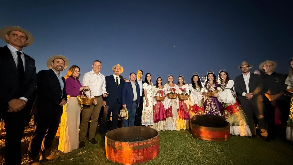 Foto posada ao ar livre, no campo, &agrave; noite. Um grupo de cerca de 15 pessoas aparece de p&eacute;, enfileirada, junto a barris de vinho. Algumas est&atilde;o com trajes formais, outras com roupas t&iacute;picas do RS.