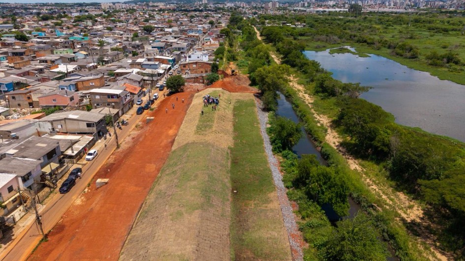 A imagem mostra uma vista a&eacute;rea de um bairro. De um lado h&aacute; muitas casas simples e pr&oacute;ximas umas das outras; do outro lado h&aacute; uma &aacute;rea verde com &aacute;gua, como um lago ou brejo. Entre a parte das casas e a &aacute;rea alagada existe um grande barranco comprido e alto de terra coberta por grama, parecido com um muro natural. Em cima desse barranco h&aacute; um pequeno grupo de pessoas reunidas. O c&eacute;u est&aacute; claro com nuvens.