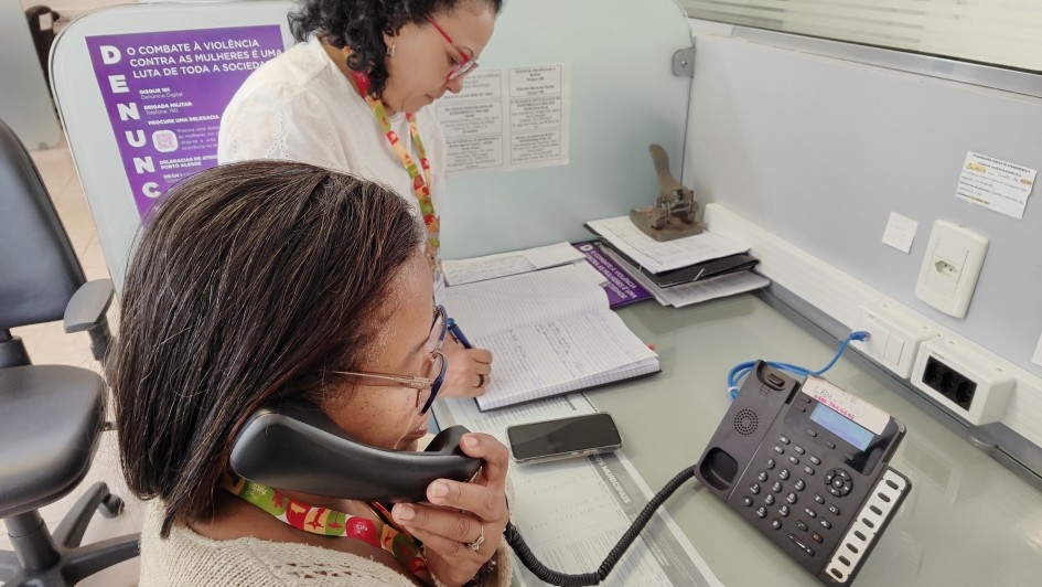 Uma fotografia colorida em plano m&eacute;dio e &acirc;ngulo levemente superior captura duas mulheres trabalhando em uma esta&ccedil;&atilde;o de atendimento de escrit&oacute;rio, focadas no servi&ccedil;o de suporte telef&ocirc;nico.

Mulher em Primeiro Plano: Uma mulher negra de cabelos escuros e lisos, usando &oacute;culos de grau e um casaco de tric&ocirc; bege. Ela segura o fone de um telefone fixo preto no ouvido esquerdo enquanto escreve em um pequeno bloco de notas com uma caneta azul.

Mulher ao Fundo: Uma mulher de pele clara e cabelos escuros cacheados, usando &oacute;culos de arma&ccedil;&atilde;o vermelha e uma blusa branca. Ela est&aacute; curvada sobre a mesa, concentrada em escrever em um grande livro de registros ou caderno aberto.

O Ambiente:

As duas trabalham em uma mesa cinza dividida por uma parti&ccedil;&atilde;o de vidro fosco.

Na lateral da divis&oacute;ria, h&aacute; um cartaz roxo e branco com a palavra "DENUNCIE" escrita verticalmente em destaque, acompanhada de textos sobre o combate &agrave; viol&ecirc;ncia contra a mulher e canais de ajuda.

Sobre a mesa, al&eacute;m dos cadernos e canetas, h&aacute; um telefone digital moderno com teclado num&eacute;rico e uma pequena tela, um smartphone e outros documentos impressos.

Contexto: A imagem retrata o atendimento da Escuta Lil&aacute;s, um servi&ccedil;o especializado de apoio a mulheres.