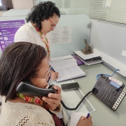 Uma fotografia colorida em plano m&eacute;dio e &acirc;ngulo levemente superior captura duas mulheres trabalhando em uma esta&ccedil;&atilde;o de atendimento de escrit&oacute;rio, focadas no servi&ccedil;o de suporte telef&ocirc;nico.

Mulher em Primeiro Plano: Uma mulher negra de cabelos escuros e lisos, usando &oacute;culos de grau e um casaco de tric&ocirc; bege. Ela segura o fone de um telefone fixo preto no ouvido esquerdo enquanto escreve em um pequeno bloco de notas com uma caneta azul.

Mulher ao Fundo: Uma mulher de pele clara e cabelos escuros cacheados, usando &oacute;culos de arma&ccedil;&atilde;o vermelha e uma blusa branca. Ela est&aacute; curvada sobre a mesa, concentrada em escrever em um grande livro de registros ou caderno aberto.

O Ambiente:

As duas trabalham em uma mesa cinza dividida por uma parti&ccedil;&atilde;o de vidro fosco.

Na lateral da divis&oacute;ria, h&aacute; um cartaz roxo e branco com a palavra "DENUNCIE" escrita verticalmente em destaque, acompanhada de textos sobre o combate &agrave; viol&ecirc;ncia contra a mulher e canais de ajuda.

Sobre a mesa, al&eacute;m dos cadernos e canetas, h&aacute; um telefone digital moderno com teclado num&eacute;rico e uma pequena tela, um smartphone e outros documentos impressos.

Contexto: A imagem retrata o atendimento da Escuta Lil&aacute;s, um servi&ccedil;o especializado de apoio a mulheres.