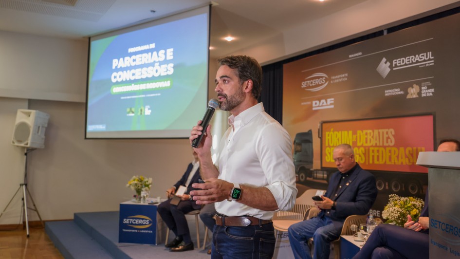 Uma fotografia colorida em plano m&eacute;dio captura o governador Eduardo Leite discursando durante um evento institucional em um audit&oacute;rio.

A&ccedil;&atilde;o Principal: O governador est&aacute; em p&eacute;, posicionado &agrave; direita do centro da imagem, falando ao p&uacute;blico. Ele segura um microfone preto com a m&atilde;o direita pr&oacute;xima ao rosto e gesticula com a m&atilde;o esquerda. Sua express&atilde;o &eacute; focada e comunicativa.

Eduardo Leite: Veste uma camisa social branca com as mangas dobradas, cal&ccedil;a jeans escura e um cinto de couro marrom. No pulso esquerdo, destaca-se um smartwatch com o visor digital aceso em verde.

O Cen&aacute;rio (Palco):

&Agrave; esquerda: Uma tela de proje&ccedil;&atilde;o exibe o t&iacute;tulo da apresenta&ccedil;&atilde;o: "PROGRAMA DE PARCERIAS E CONCESS&Otilde;ES - CONCESS&Otilde;ES DE RODOVIAS" sobre um fundo verde e azul.

Ao fundo: H&aacute; um painel (backdrop) com as logomarcas da SETCERGS, FEDERASUL, DAF e do Governo do Estado do Rio Grande do Sul. O painel tamb&eacute;m traz o t&iacute;tulo: "F&Oacute;RUM DE DEBATES SETCERGS | FEDERASUL".

Participantes: Dois homens est&atilde;o sentados em cadeiras beges atr&aacute;s do governador, ouvindo o discurso. &Agrave; direita, v&ecirc;-se parte de um p&uacute;lpito met&aacute;lico com o adesivo da SETCERGS.

O Ambiente: O audit&oacute;rio possui ilumina&ccedil;&atilde;o quente vinda de spots no teto, piso de madeira (parquet) e uma caixa de som branca montada em um trip&eacute; &agrave; esquerda.